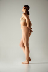 Side view full-length image of young girl with with, smooth, slim body, buttocks. Model posing in underwear against grey studio background. Concept of body and health care, female beauty, wellness