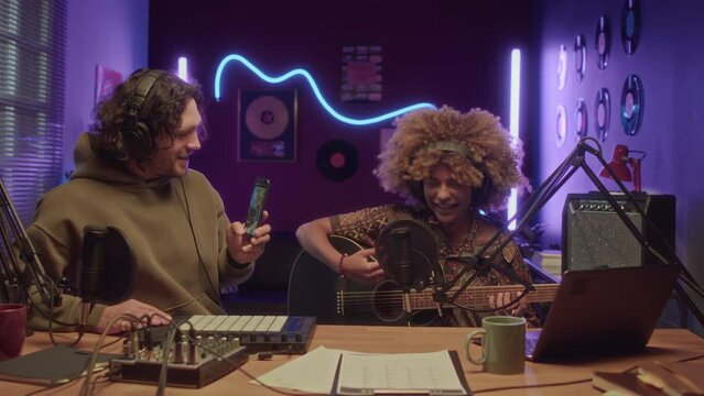 UGC footage of Caucasian man holding smartphone and filming African American female friend who singing song to guitar in neon lit home recording studio at night