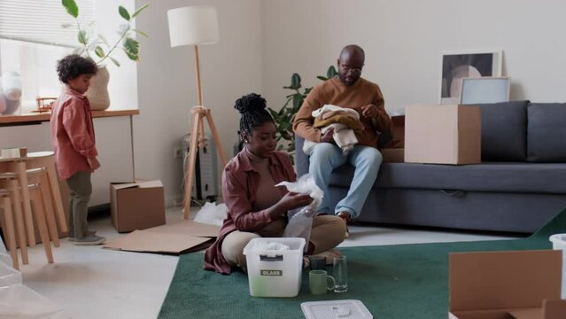 African American family couple packing glasses and clothing into boxes with help of little son while getting ready for moving in new apartment