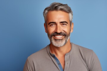 Portrait of handsome mature man with grey hair and beard smiling at camera against blue background - obrazy, fototapety, plakaty
