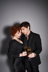 charming woman in black dress hugging her handsome boyfriend on gray backdrop, champagne in hands