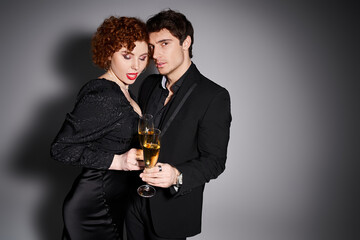 charming woman in black dress hugging her handsome boyfriend on gray backdrop, champagne in hands