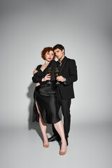 beautiful woman in black dress hugging her handsome boyfriend on gray backdrop, champagne in hands