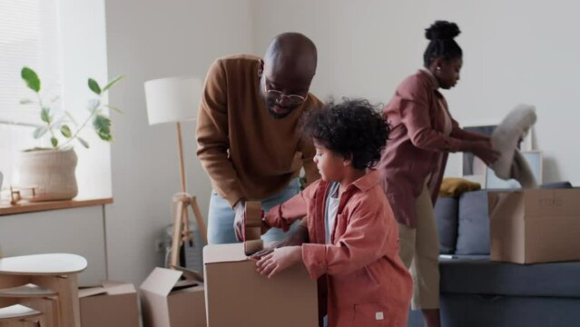 African American man sealing cardboard box with help of little son while packing belongings before moving with family