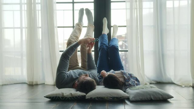 young lovers laying on floor with feet up on window romantic chatting and smiling in living room. beloved young man and woman hold mobile phone, using online app or taking selfie photos