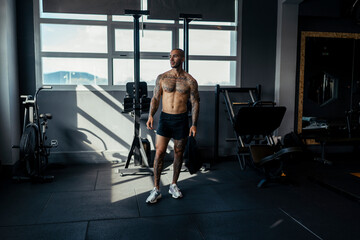 Chico joven tatuado y musculoso posando en gimnasio sin camiseta y con ropa moderna