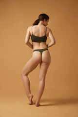 Full-length image of brunette young girl with slim, healthy, relief body, posing in cotton underwear against beige studio background. Concept of body and health care, female beauty, wellness