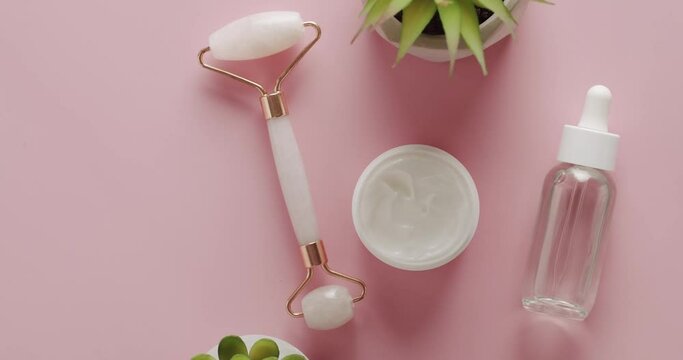 Top view on jade roller for face massage, open jar with hydrating cream and rejuvenation serum in dropper bottle on pink background. Cosmetic products for face lifting and anti wrinkles procedures.