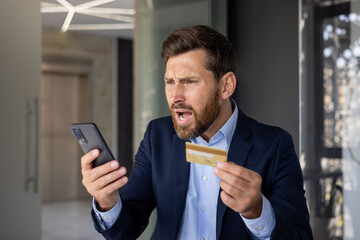 Close-up photo of angry young businessman in suit standing in office, holding golden credit card in hand and looking disappointedly at mobile phone screen - obrazy, fototapety, plakaty