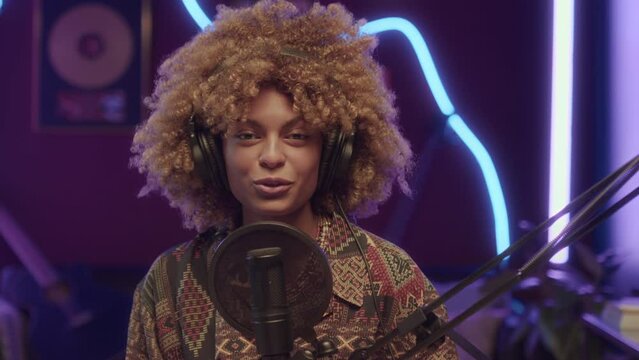 Chest up UCC of cheerful African American girl talking into microphone while recording online program or video for vlog at home studio with neon string lights at night