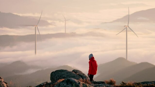 Young woman stand against landscape with wind turbines on background. Girl in orange jacket running up on top of mountain summit at sunset, raises arms into air, happy and drunk on life and happiness