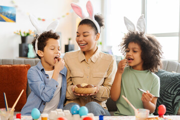 Portrait happy latin family eating chocolate eggs at home. Smiling mother and kids wearing bunny...