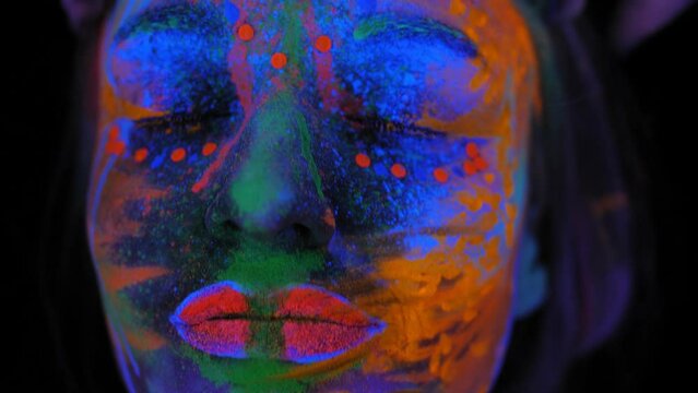 A close-up of a girl's face with an ultraviolet pattern, she moves her hands in the light of a fluorescent lamp, neon paint glows on her face. A female model in neon light.