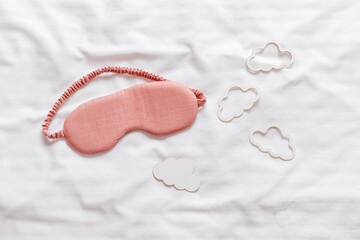 Sleeping mask pink color and clouds shapes on bed white bedclothes background. Eye cover for best sleep. Creative Concept of dream well and good night, comfort rest at night. Top view