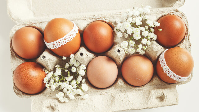 Brown Easter eggs decorated lace and white gypsophila flowers in carton on white background. Easter celebration concept. Top view photo of festive food of painted chicken egg retro toned