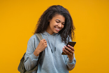 Half-length studio portrait of beautiful casually dressed girl with perfect charming smile texting message via her mobile phone isolated on bright colored yellow background