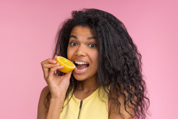 Studio shot of cute and funny african american girl posing over pastel pink background pretending to bite a lemon cut in a half - 745787229