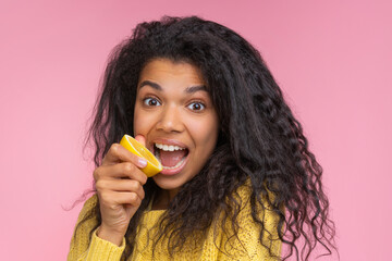 Close up portrait of cute and funny african american girl posing over pastel pink background pretending to bite a lemon cut in a half - 745787291