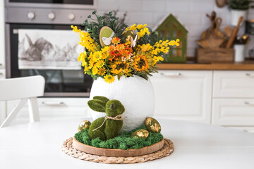 A spring bouquet in an egg, Easter bunnies and eggs with a golden pattern on the table. In the background is a white Scandinavian-style kitchen. A beautiful greeting card. Easter decor in the house.