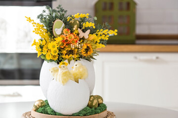 A spring bouquet, yellow little chickens in a large egg and Easter eggs with a golden pattern on the table. In the background is a white Scandinavian-style kitchen. Easter decor in the house.