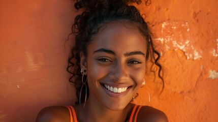 A young woman with curly hair wearing hoop earrings smiling against an orange textured wall.