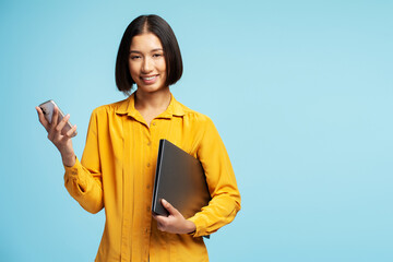 Portrait of smiling woman wearing yellow shirt holding mobile phone and laptop looking at camera