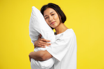 Cute beautiful smiling Asian woman holding white pillow standing isolated on yellow background