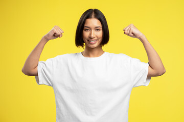 Smiling strong asian girl showing biceps, muscles standing isolated on yellow background