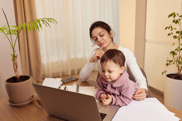 tired woman working on laptop at home with her little baby girl. Child makes noise and disturb mother at work.