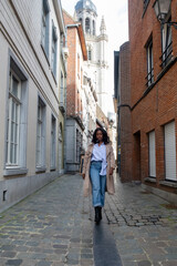 Fototapeta na wymiar A young African woman is captured mid-stride walking down a narrow cobblestone street in an old European town. The historic charm of the town is exemplified by the traditional architecture and the
