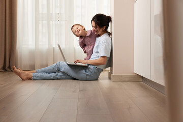 happy casual woman sitting on the floor and working on laptop with her little baby girl at home