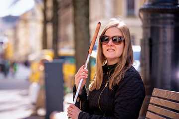 A visually impaired woman holding a white cane and sitting on a bench in the city