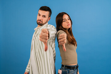Couple man and woman looking at camera with sad expression and thumbs down. Portrait of young male and female doing disapprove symbol and unhappy expression on blue background studio