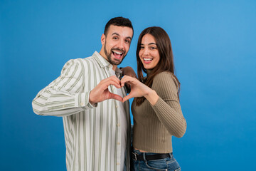 Young couple, male and female, looking at camera doing heart shape symbol with their hands and smiling at camera in blue background studio copy space