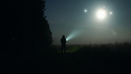 A mysterious figure with torch looking at glowing UFO lights in the sky. On a spooky misty night in the countryside.