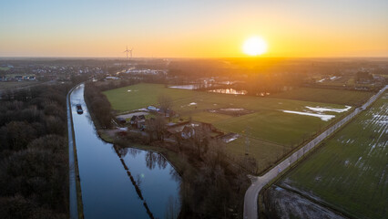 This image offers an aerial view of a serene rural scene just as the day begins, with the golden sunrise casting its warm light across the landscape. A canal winds through the scene, reflecting the