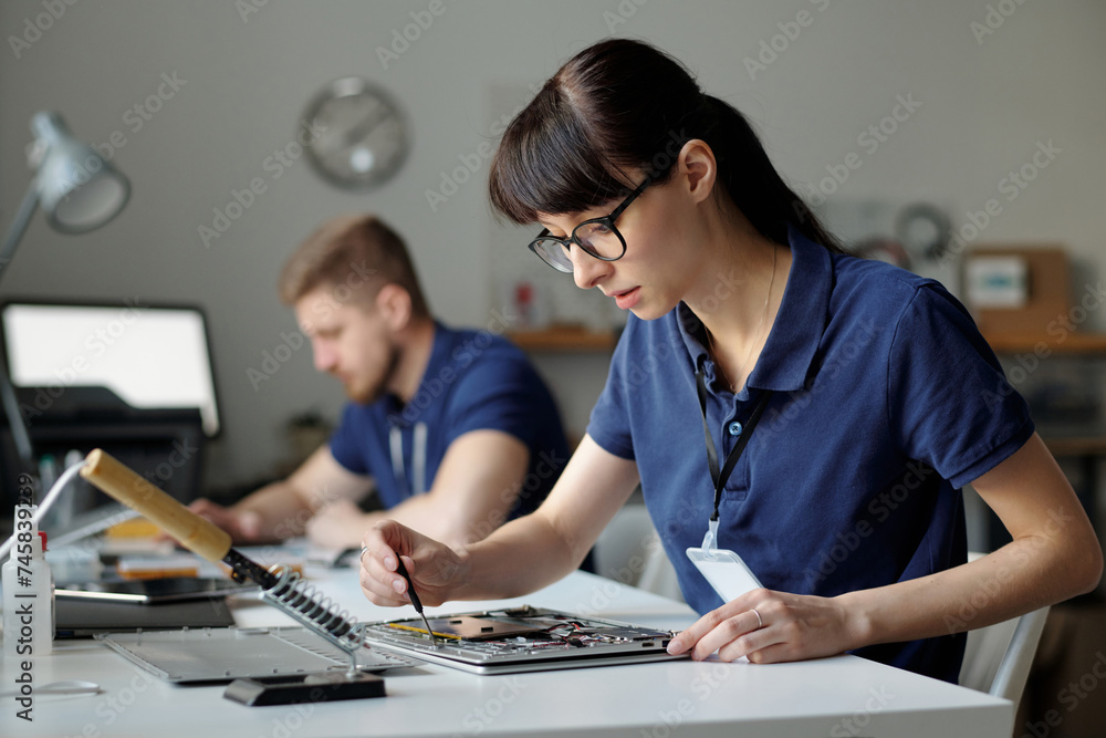 Poster Pretty technical service worker fixing tiny details of disassembled laptop while sitting by workplace in front of camera against colleague