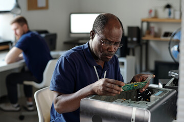 Experienced African American repairman in eyeglasses looking at computer motherboard and...