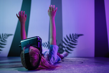Child girl with virtual reality glasses laying on the VR room floor, lit by fluorescent light effects treetop grow simulation. Science and children concepts. - obrazy, fototapety, plakaty
