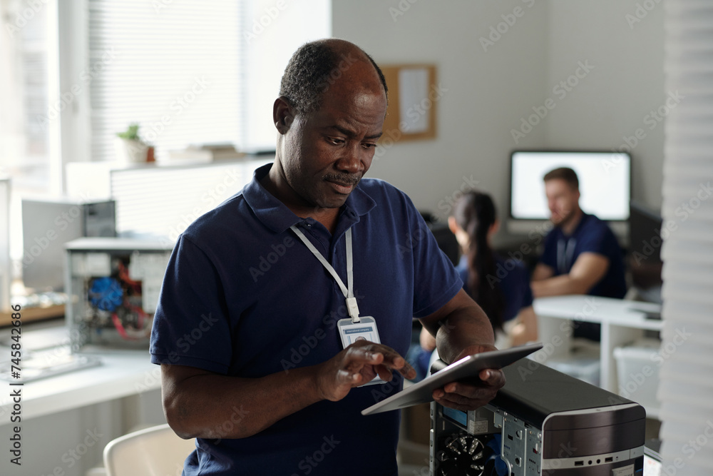 Poster Mature engineer or technician of repair center using tablet while standing by disassembled computer processor and reading online manual
