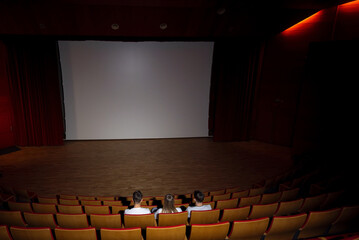Three friends, a teenage girl and two boys smiling, watching a movie in an empty cinema with red...