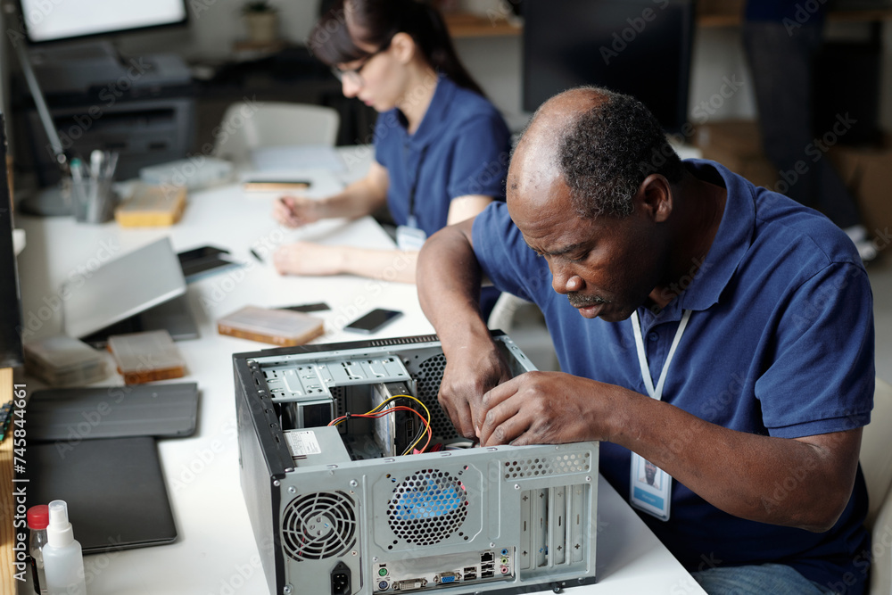 Poster professional technician sitting by desk in repair service office and bending over disassembled compu