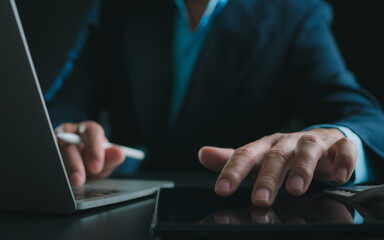 Businessman in black suit working on laptop computer and tablet, Hand Hands touch on tablet at office with dark background, Online working, Close up, Copy space.