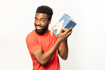 Happy African American man in red shirt, holding a gift box with a bow and smiling, isolated on a white backdrop. Perfect for expressing joy and celebration. - 745848084