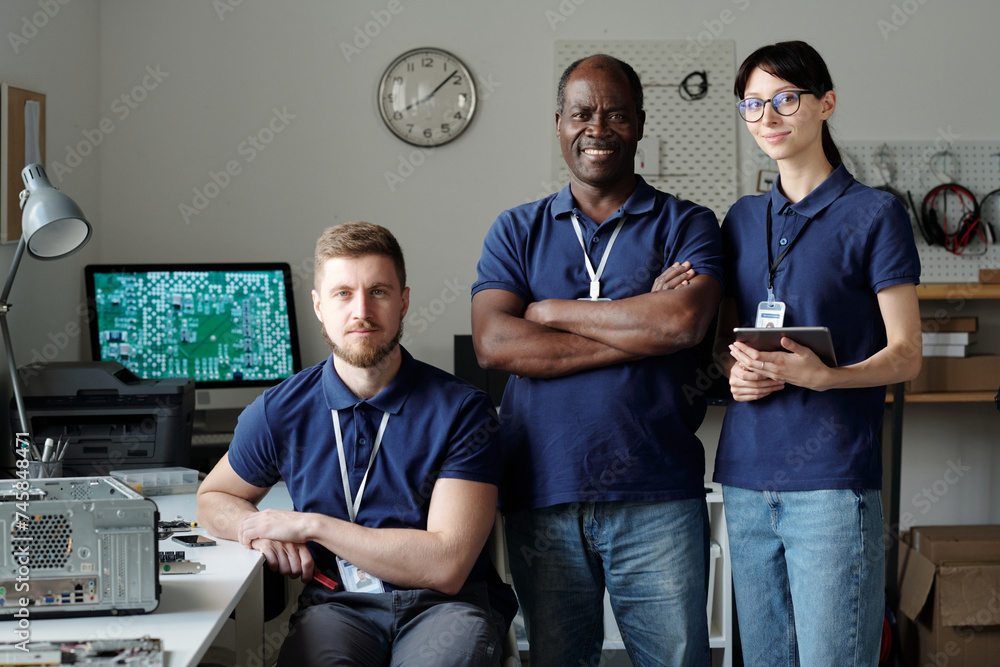 Poster Team of successful intercultural technicians in blue uniform looking at you while standing in front of camera in repair service office