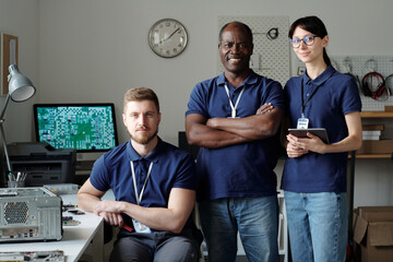Team of successful intercultural technicians in blue uniform looking at you while standing in front...