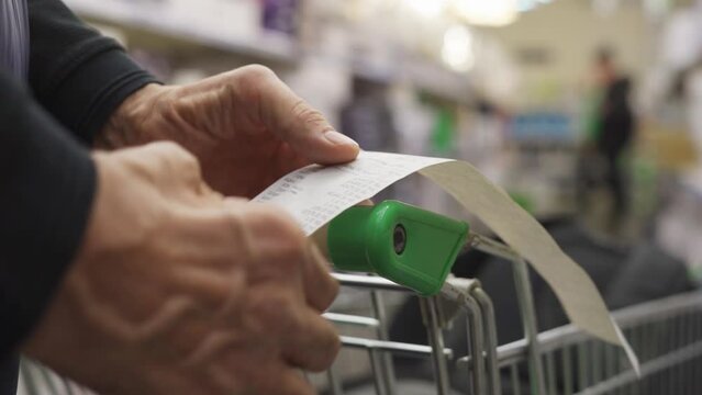The hands of an unrecognizable older man hold a sales receipt next to a grocery cart in a supermarket.