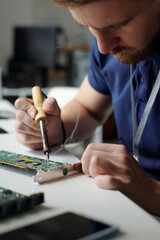 Young male engineer of maintenance service sitting by desk in front of camera and repairing computer motherboard with electric handtool