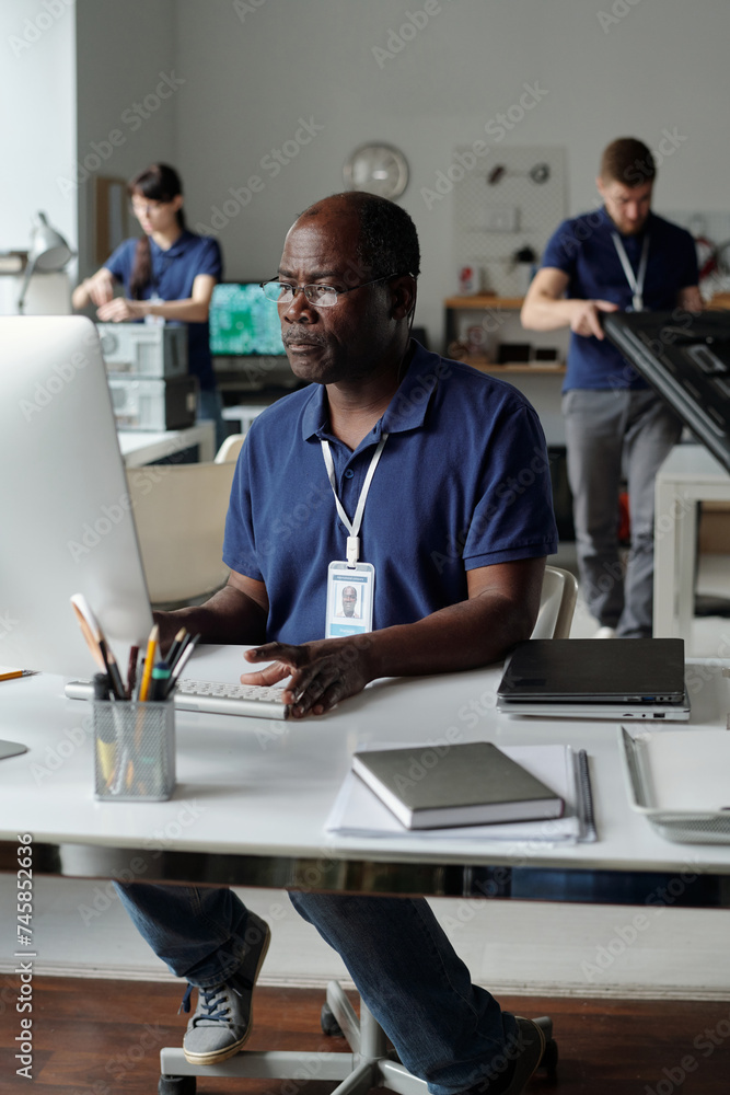 Poster experienced male engineer of repair service center looking at screen of desktop computer while sitti