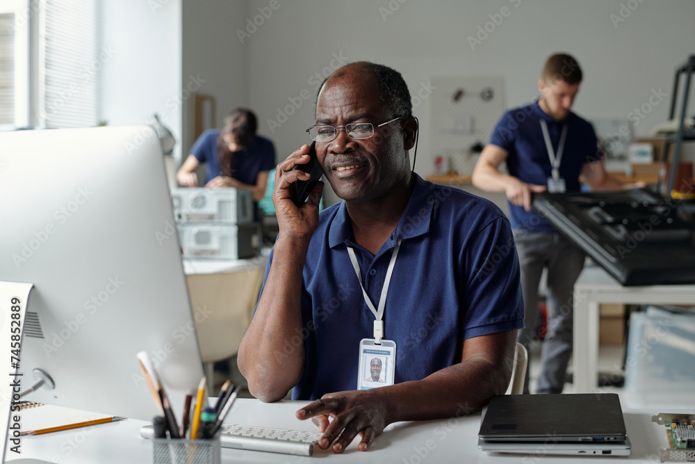 Poster Experienced technician of repair service looking at computer screen while sitting by workplace and speaking to client on smartphone